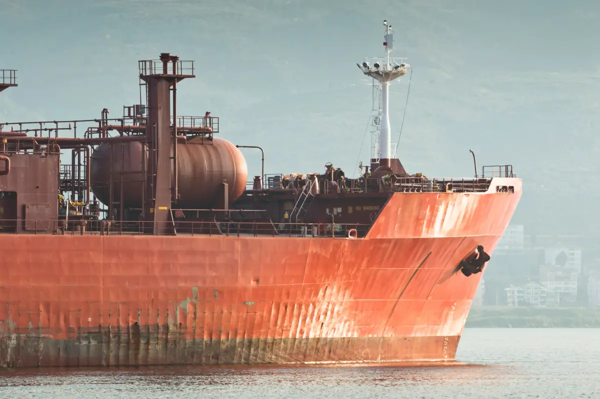 A close-up view of a large rusty industrial ship docked near a coastal city.