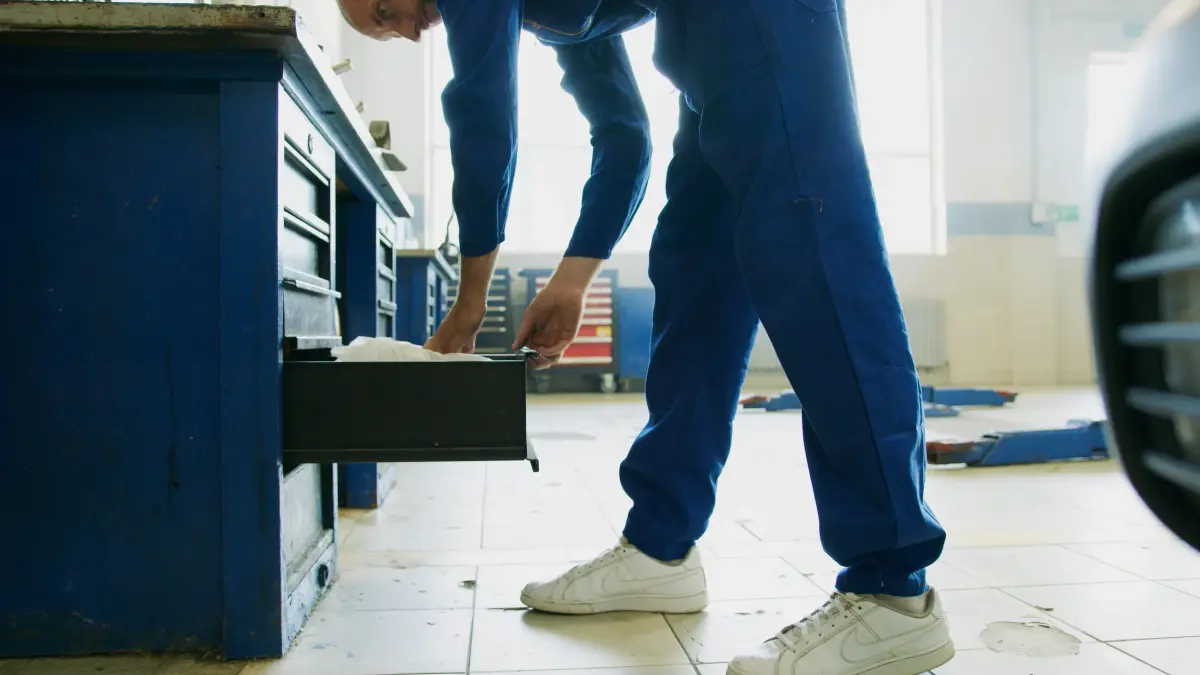Mechanic in blue coveralls opens a toolbox drawer in a sunlit workshop.