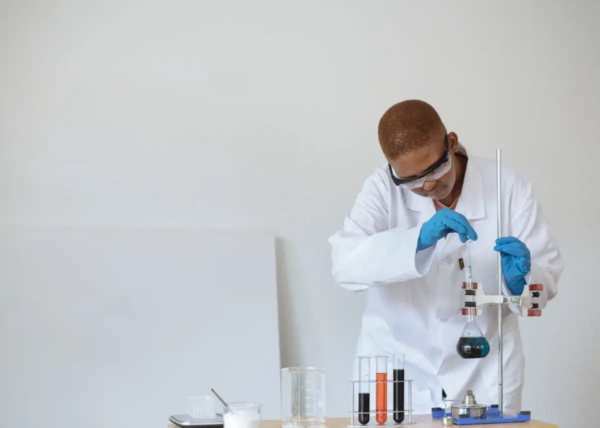 Focused African American schoolboy in safety glasses and uniform examining flask with dark liquid while conducting chemical experiment in classroom