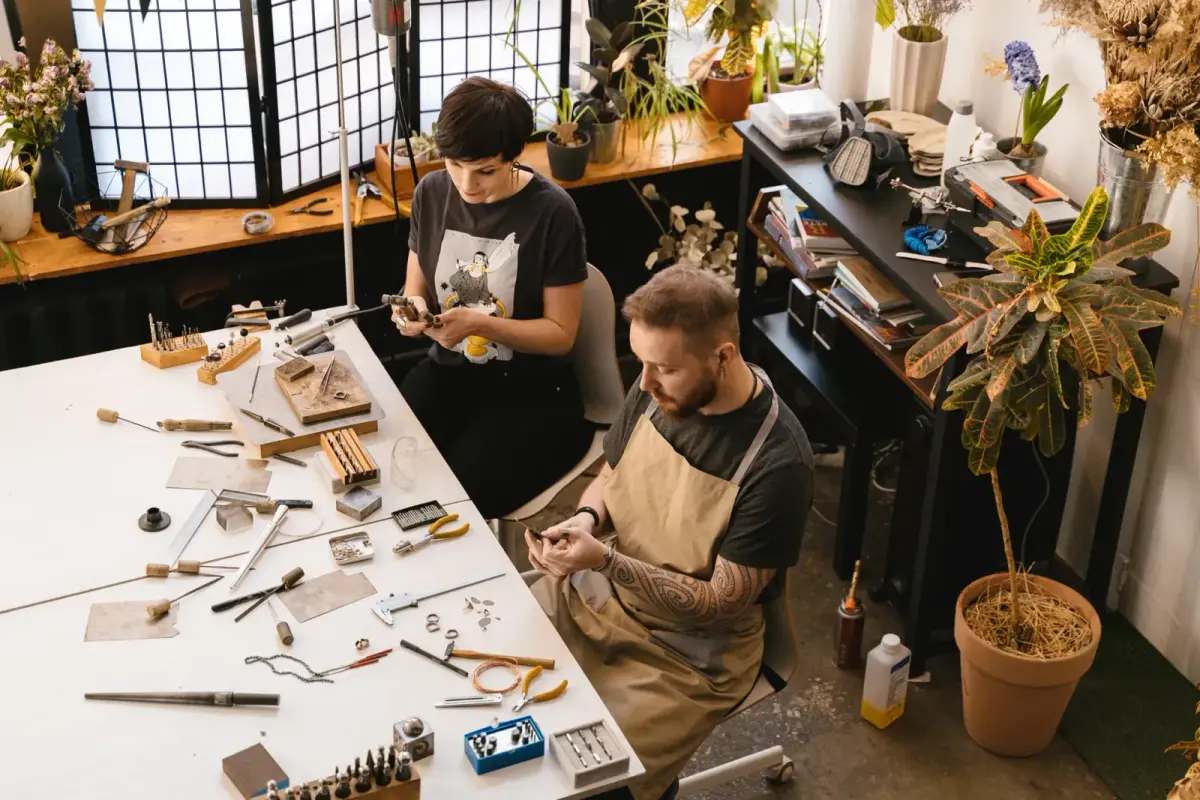 Two artisans working with tools and materials in a cozy, plant-filled workshop.