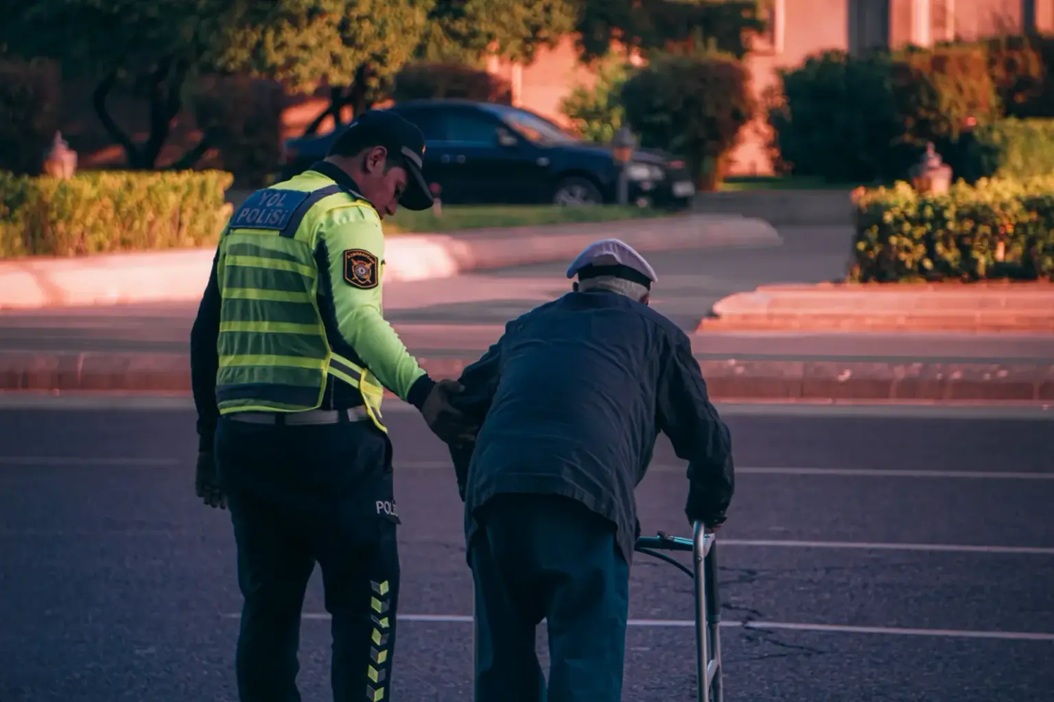 A police officer helps an elderly man with a walker cross the street at sunset.